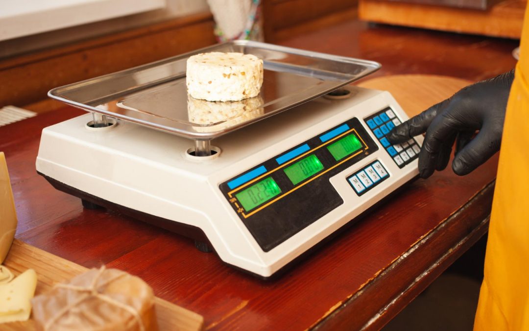 A small round cheese is being weighed on an industrial scale by a person wearing a black glove.